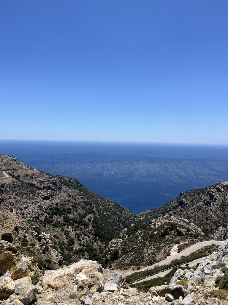 A winding road through dry Greek hills descending toward the Aegean sea