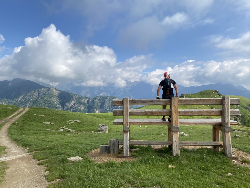 A person resting against a wooden bench on a high alpine meadow, looking out over distant mountains.